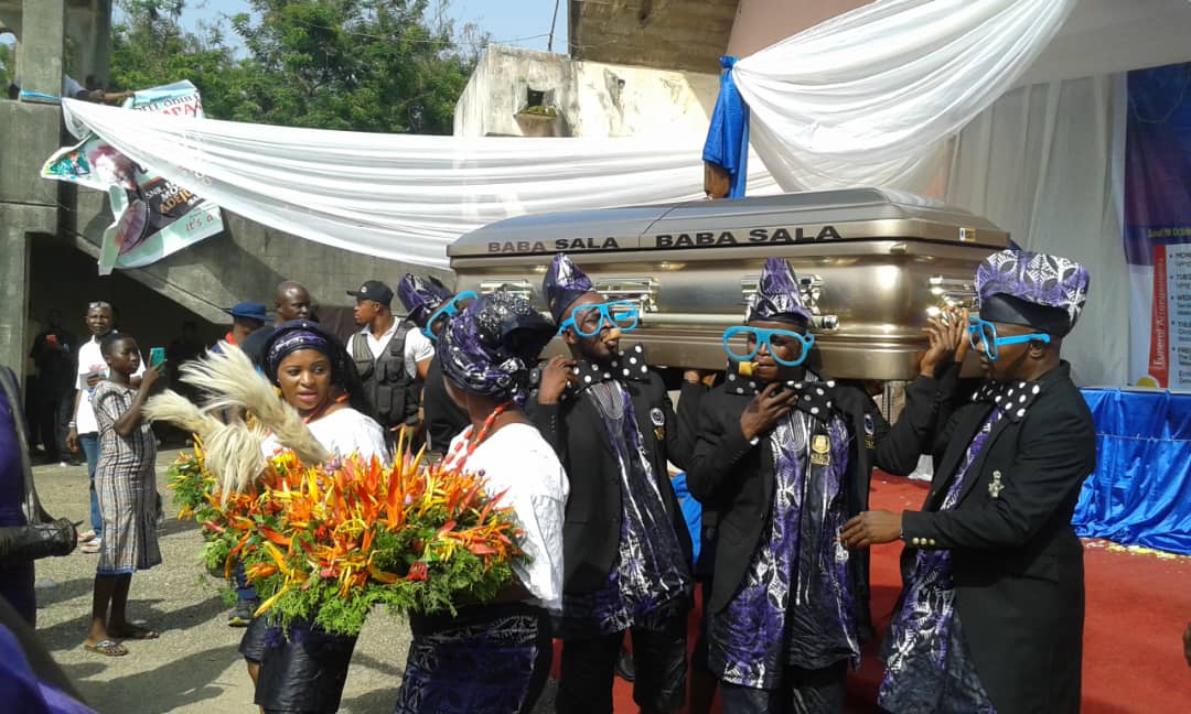 Baba Sala's life being celebrated with his trademark goggle and tie as his casket arrives for the lying in state at the Cultural Centre in Ibadan